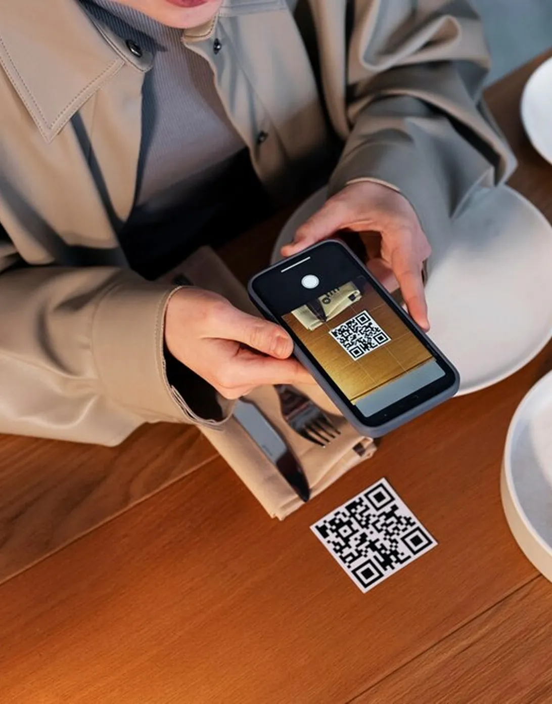 Person holding a smartphone scanning a QR code placed on a wooden table next to a napkin with cutlery and two empty white plates.