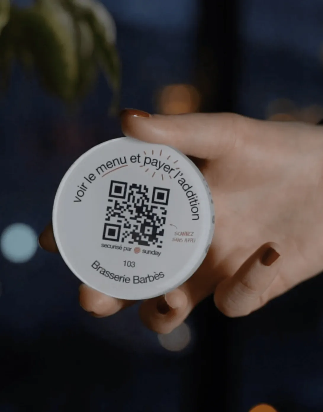 Hand holding a round token with a QR code and French text directing to view the menu and pay the bill at Brasserie Barbès.