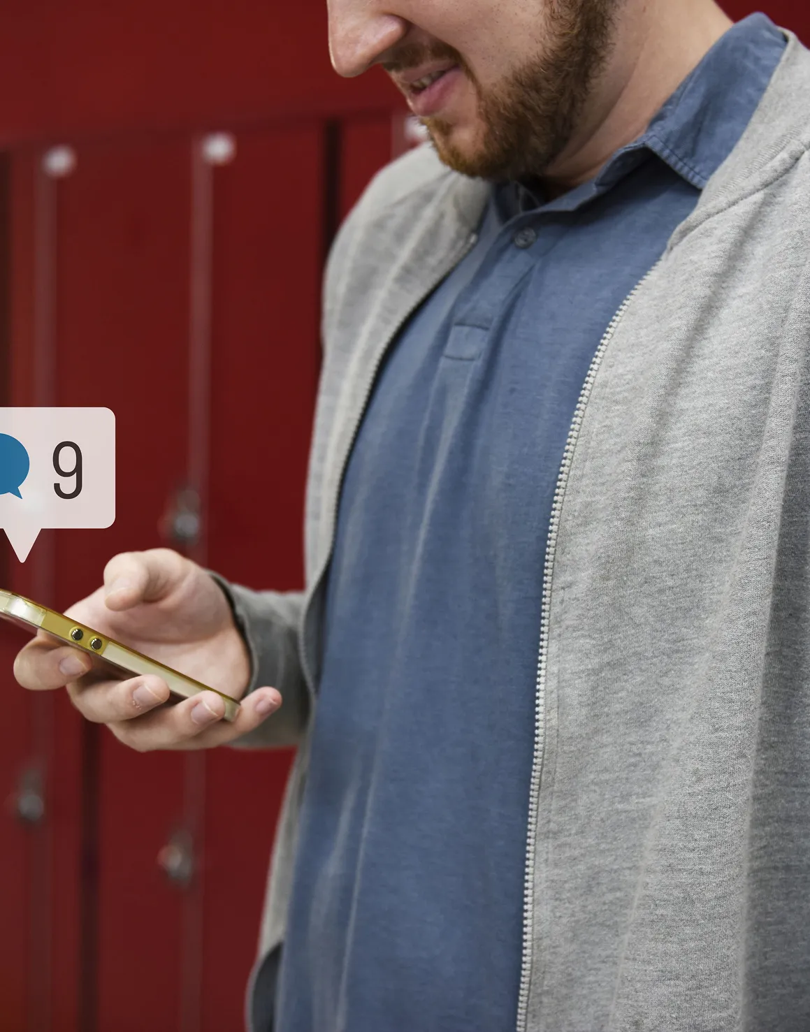 Man with beard wearing a gray hoodie and blue shirt looking at a gold smartphone with a notification bubble showing 9 messages.